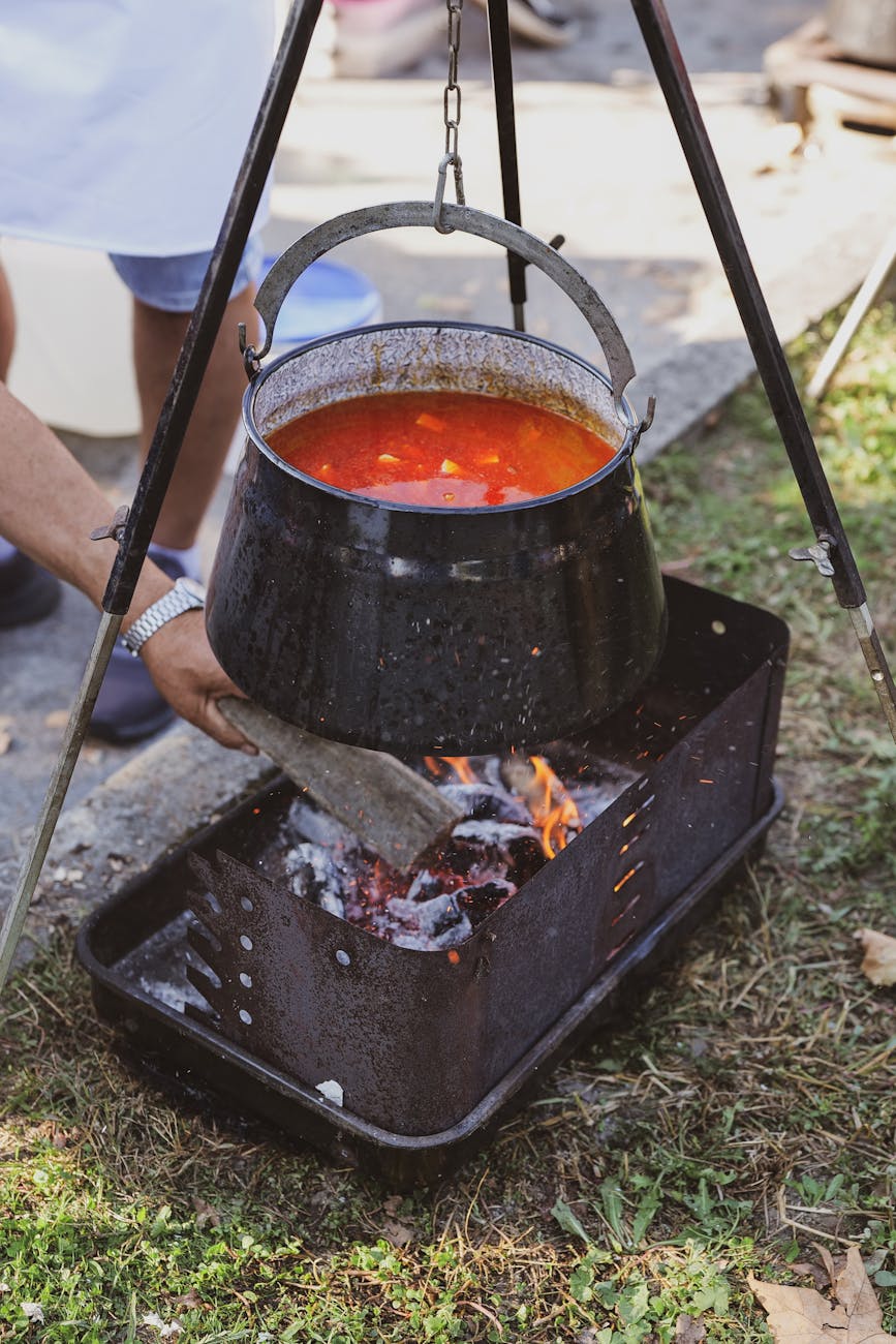 a man putting a piece of wood to a fireplace with a hanging pot of soup